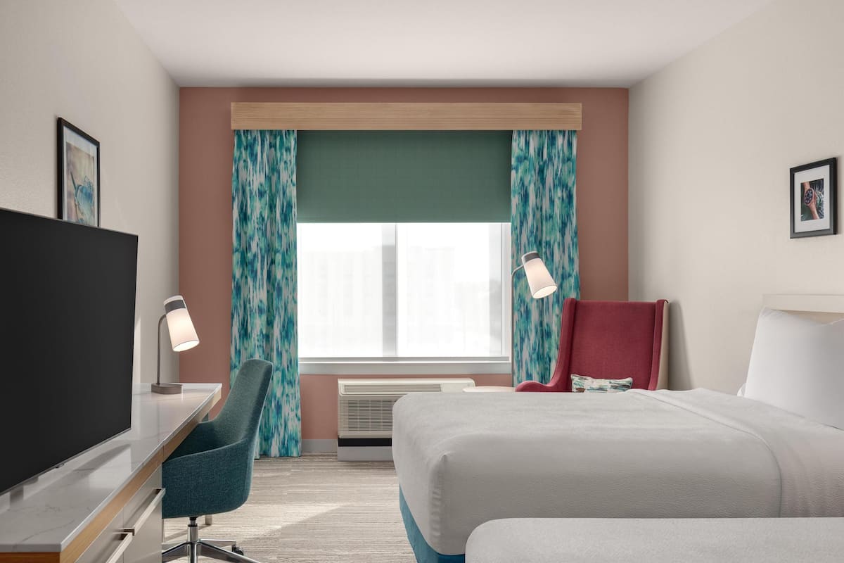Una habitación de hotel con cama, escritorio con televisor, silla, ventana con cortinas y lámpara.
