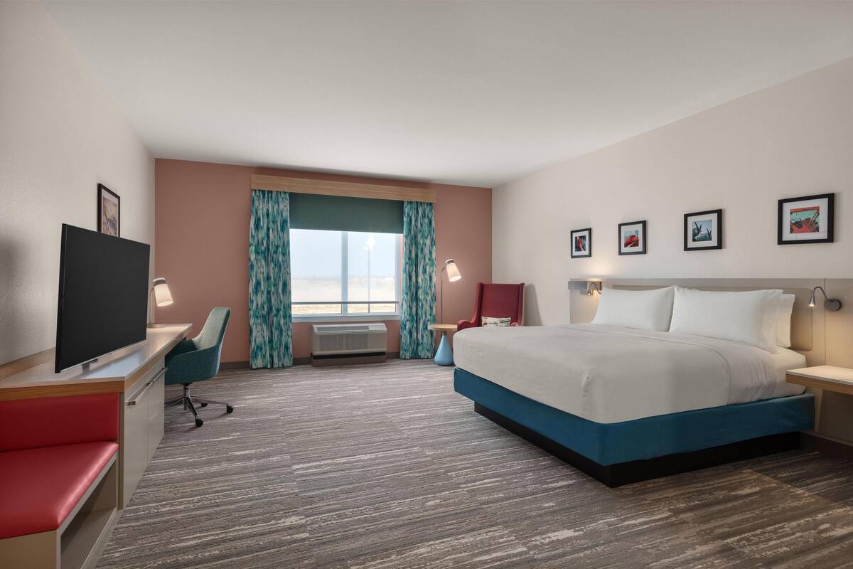 Habitación de hotel con cama, escritorio con televisor, silla, ventana con cortinas y pared decorada con cuadros.
