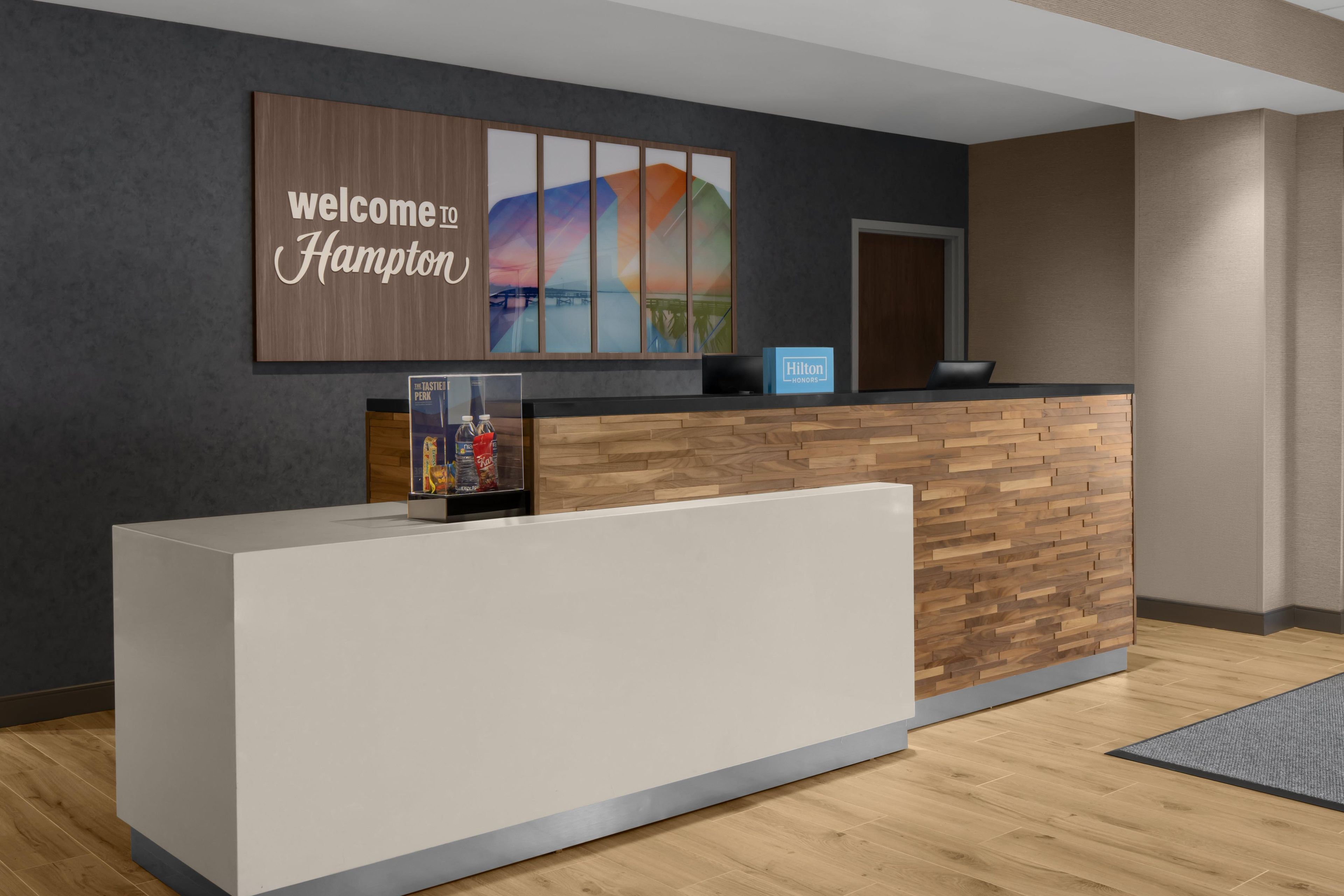 A reception desk with a 'Welcome Hampton' sign, a framed cityscape artwork, and a 'Hilton Honors' sign.