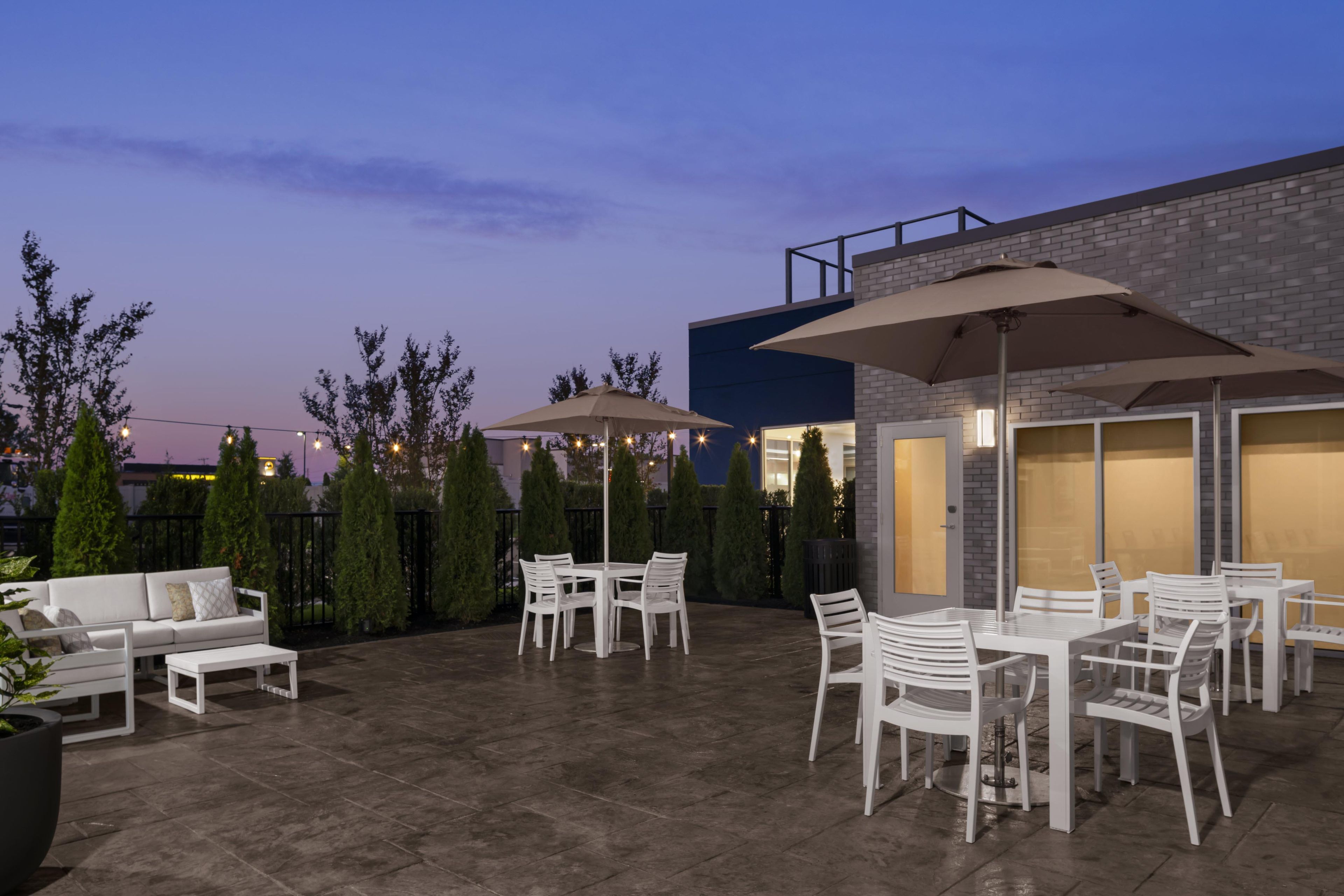 An outdoor patio area with a white table and chairs, two umbrellas, and a modern building in the background.