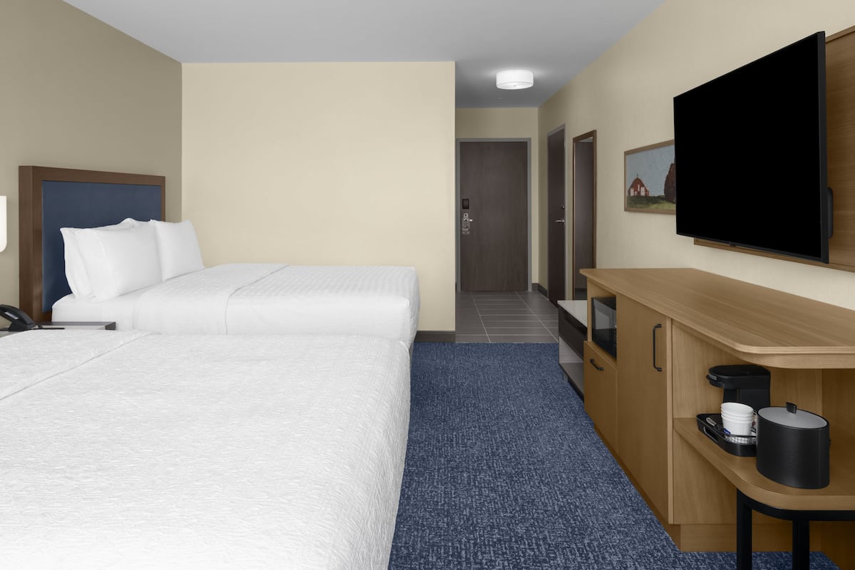 Habitación de hotel con dos camas, televisión, escritorio con cafetera y puerta que da a un pasillo.