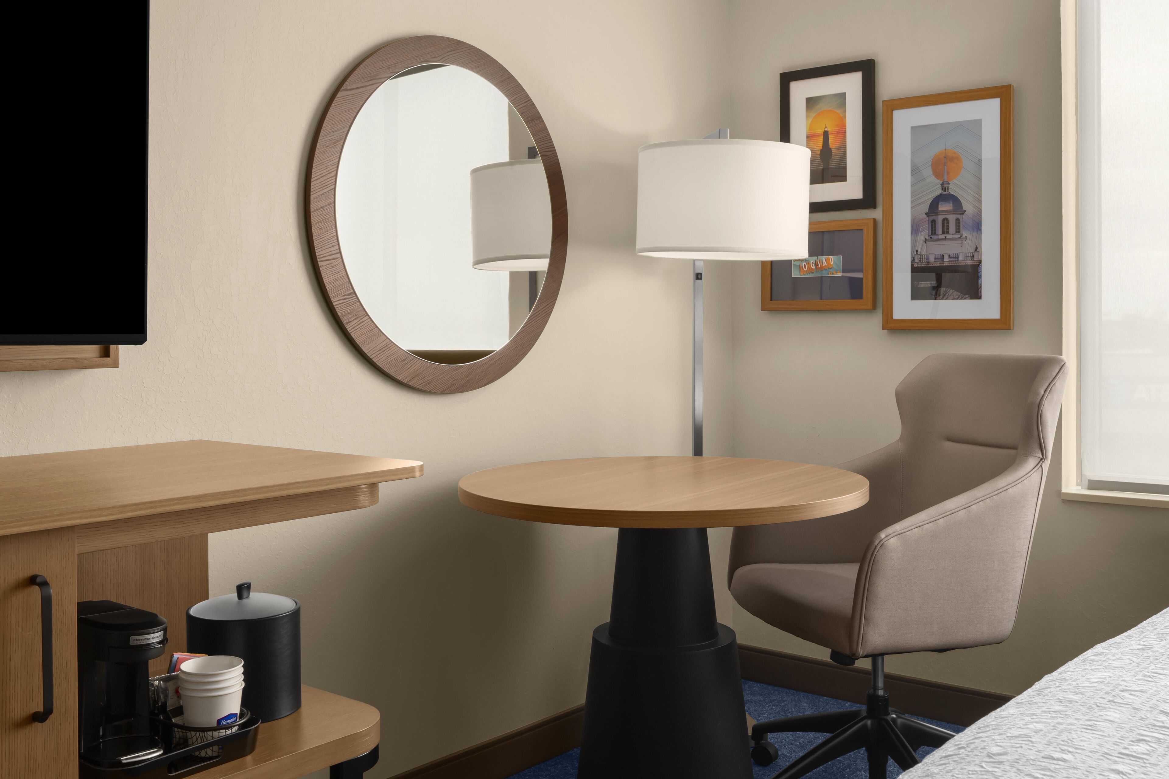 Habitación de hotel con un escritorio de madera, una mesa redonda, una silla, cafetera, lámpara y cuadros en la pared.
