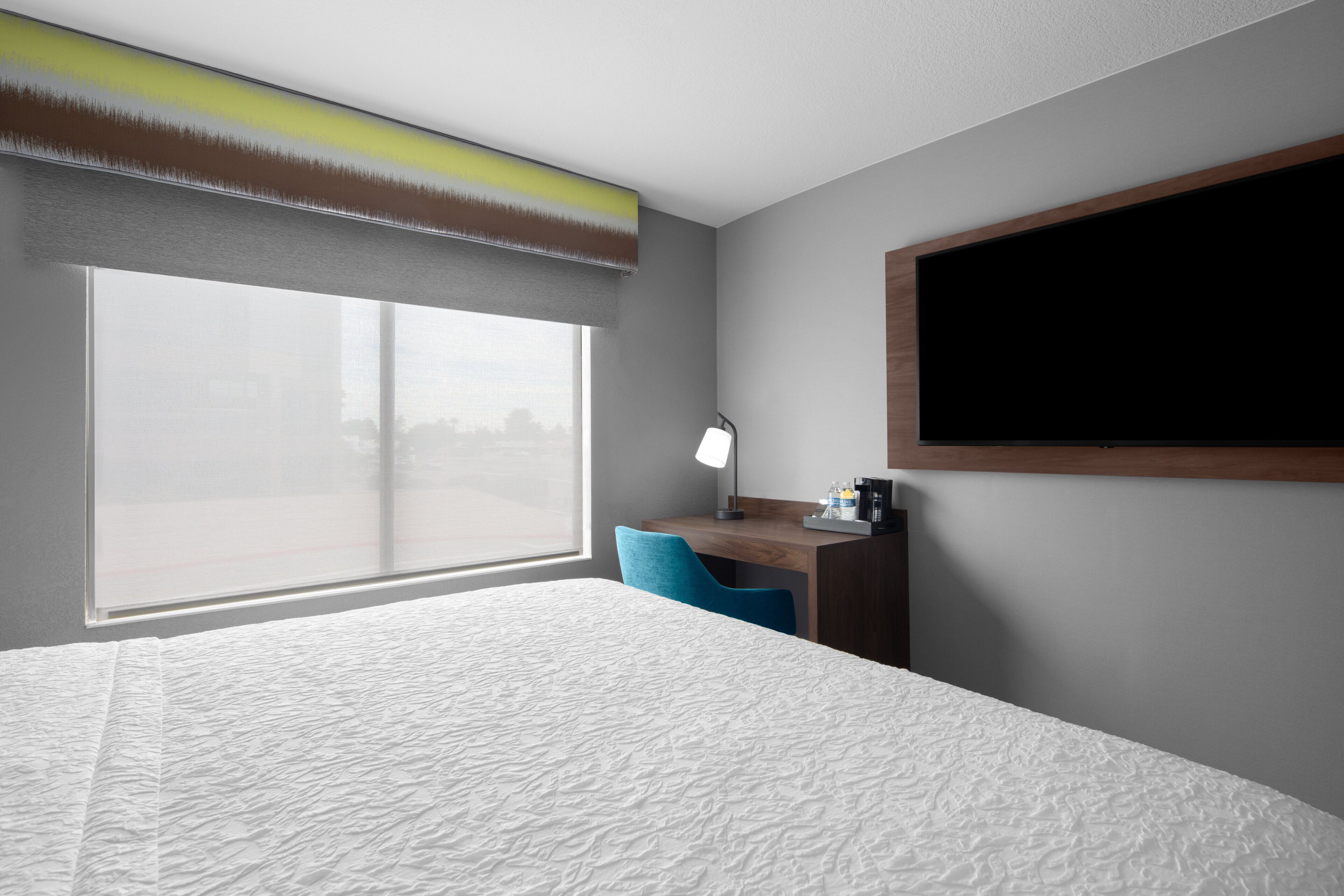 Een hotelkamer met een groot raam, een bureau met een lamp en een bed met een geribbelde matras.