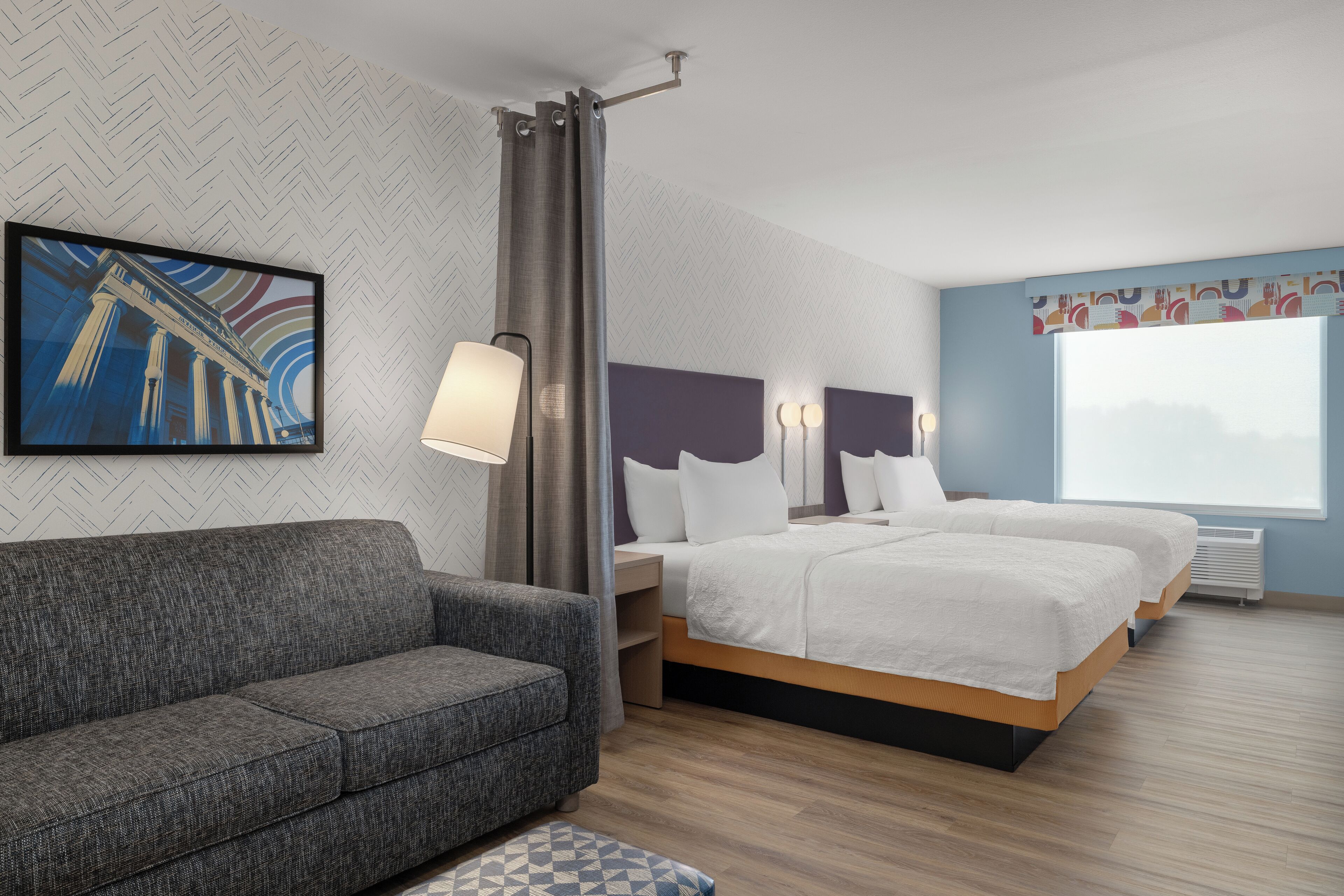 Una habitación de hotel moderna con un sofá gris, una cama con ropa de cama blanca, un ventanal grande y una obra de arte colgada en la pared.