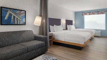 A modern hotel room with a grey sofa, a bed with white bedding, a large window, and a wall-mounted artwork.