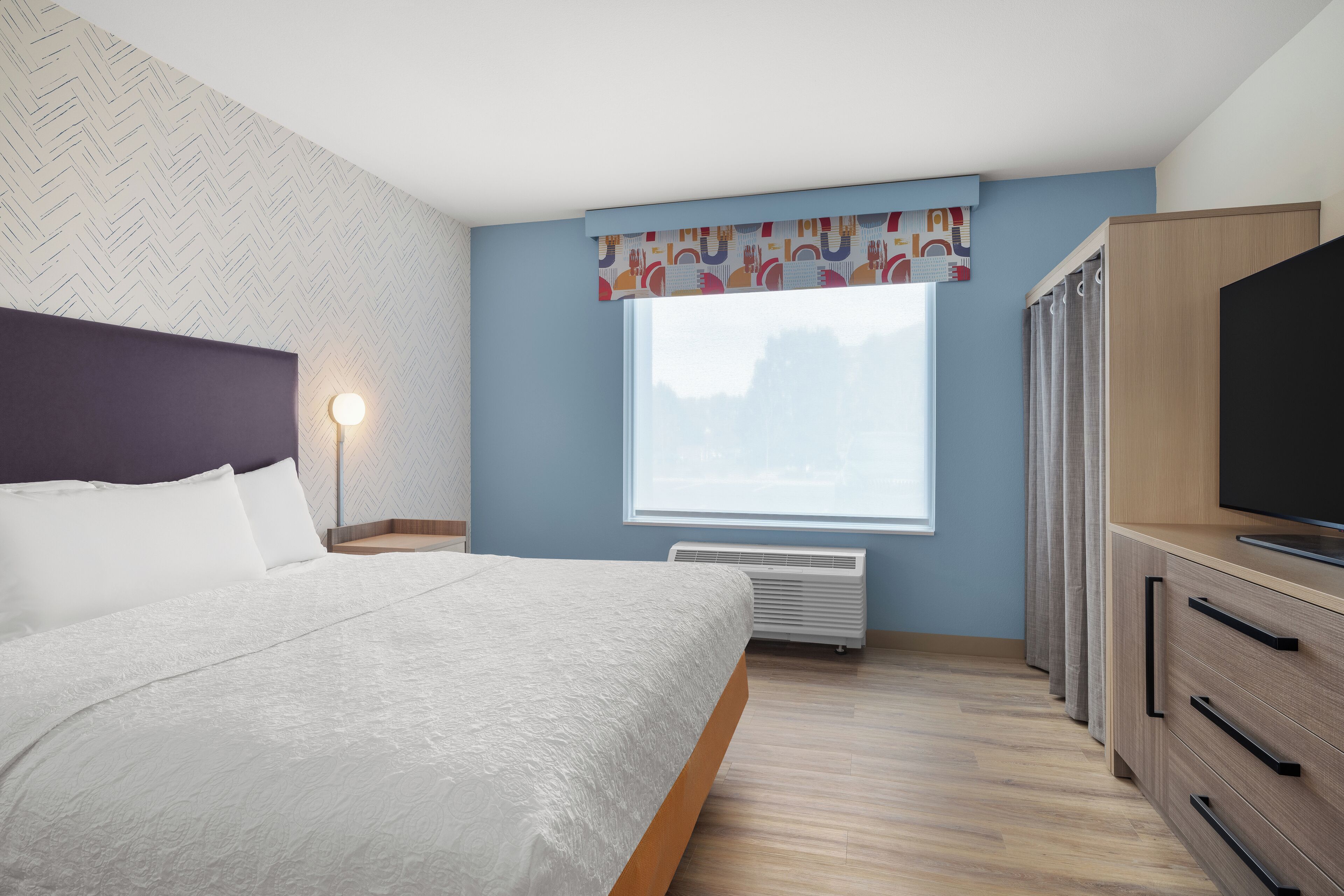 A bedroom with a bed, a bedside lamp, a window with curtains, a TV on a wooden cabinet, and a wall with a herringbone pattern.