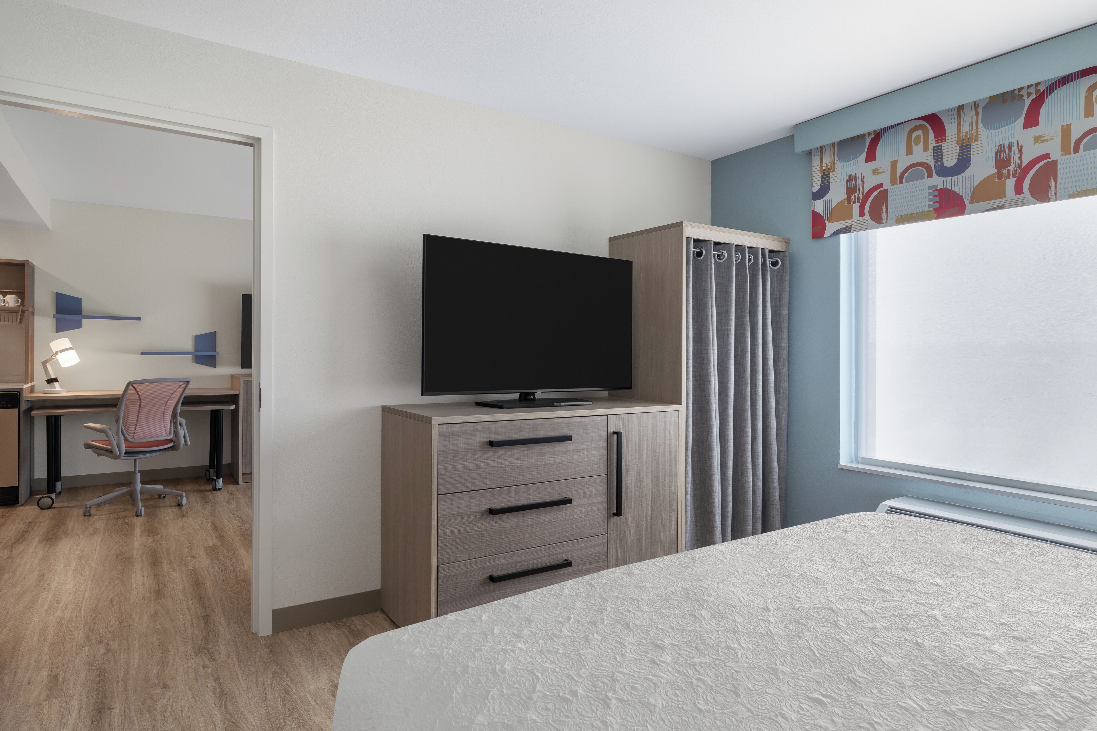 A hotel room with a bed, a desk with a chair, a television on a cabinet, and a window with curtains.