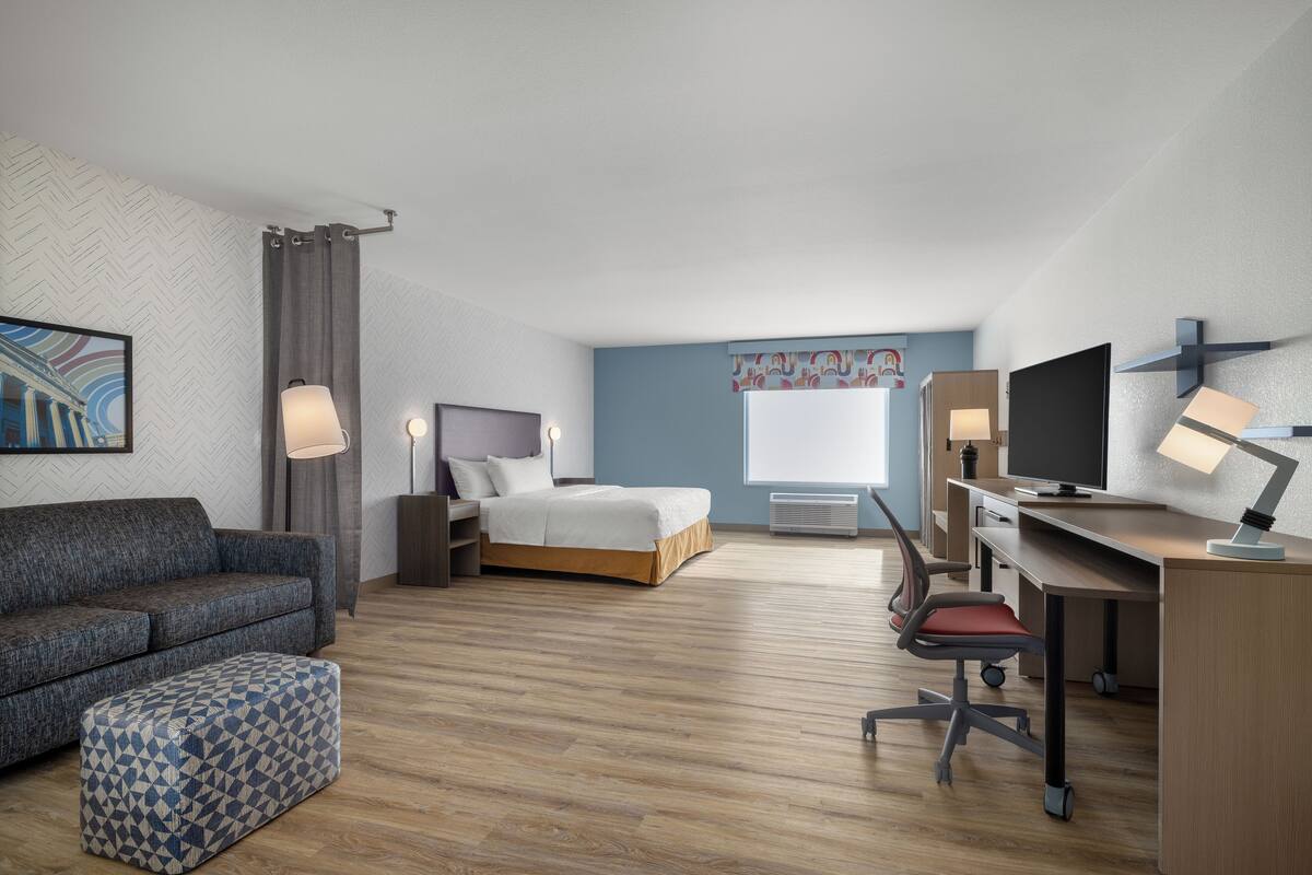 A hotel room with a bed, a desk with a computer, a TV, a sofa, and a patterned ottoman.