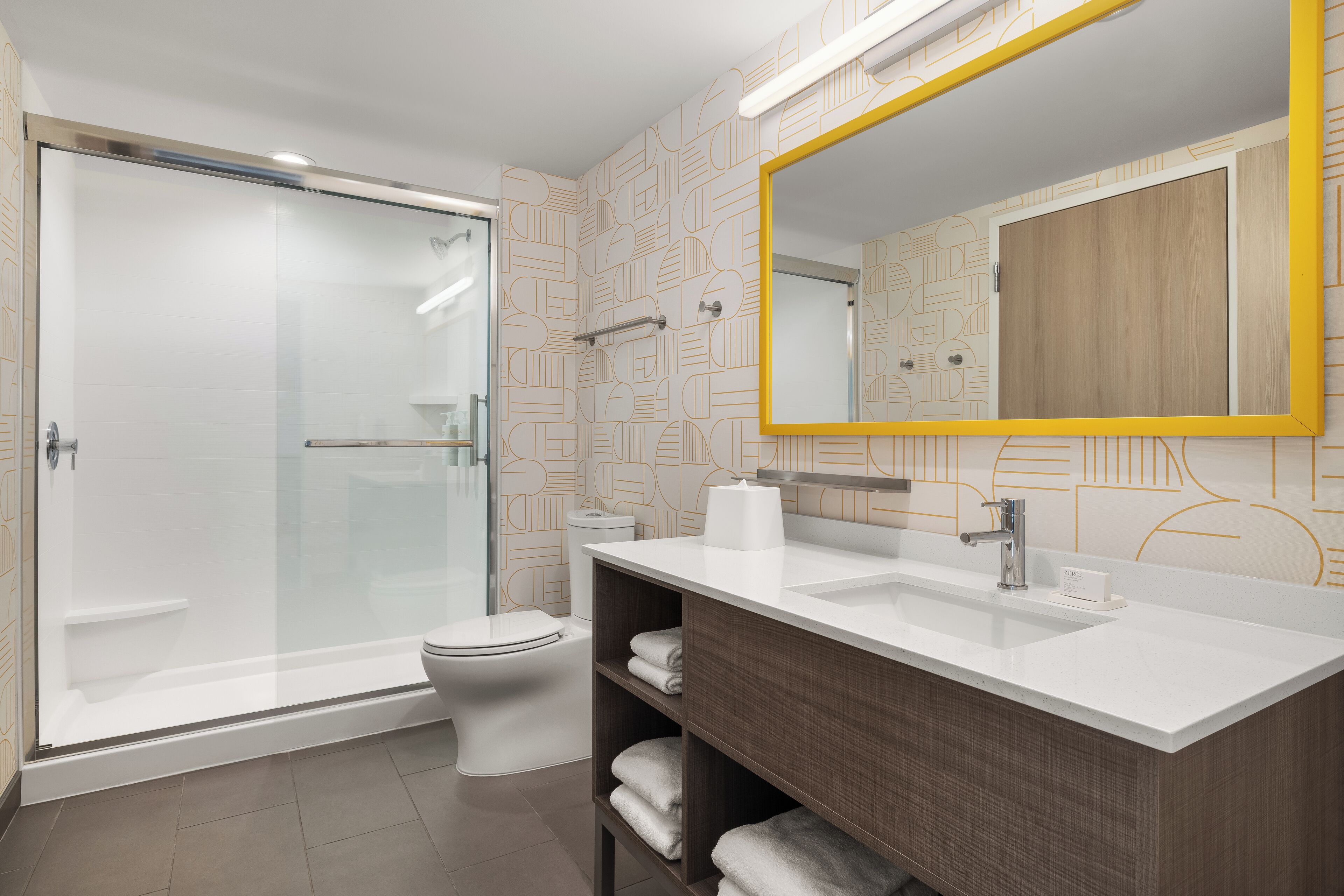 A modern bathroom with a glass shower enclosure, a toilet, a sink with a wooden vanity, and a mirror with a yellow frame.