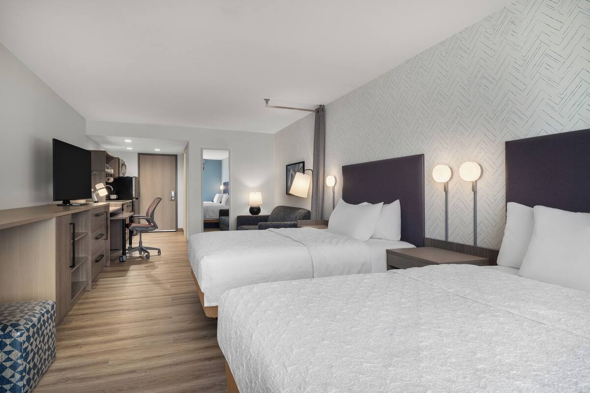 A hotel room with two beds, a desk, a chair, a TV, and a bathroom visible in the background.