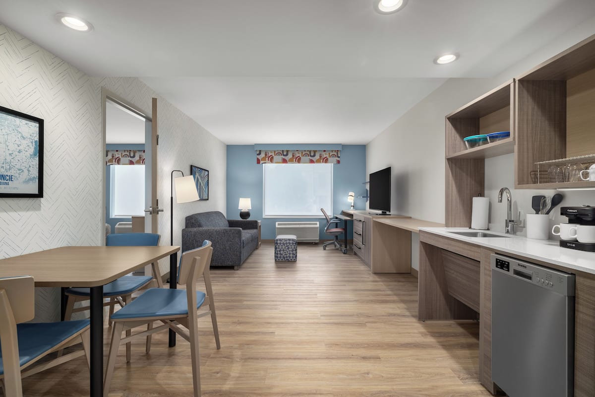A compact kitchen with a dishwasher, a dining area with a table and chairs, a sofa, and a window with curtains.