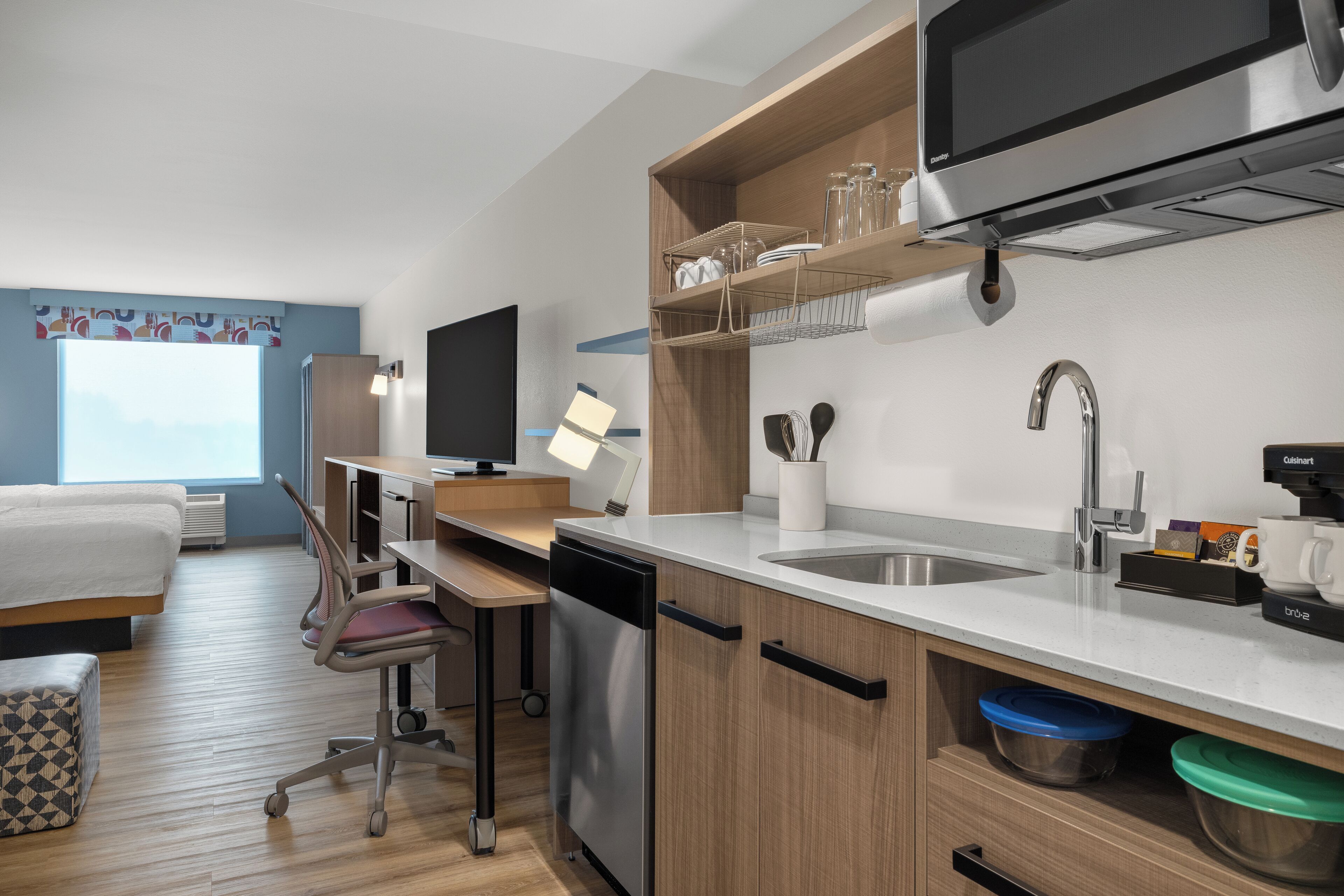 A modern kitchen with a dishwasher, sink, and built-in microwave. There is a dining area with a table and chairs, and a TV mounted on the wall.