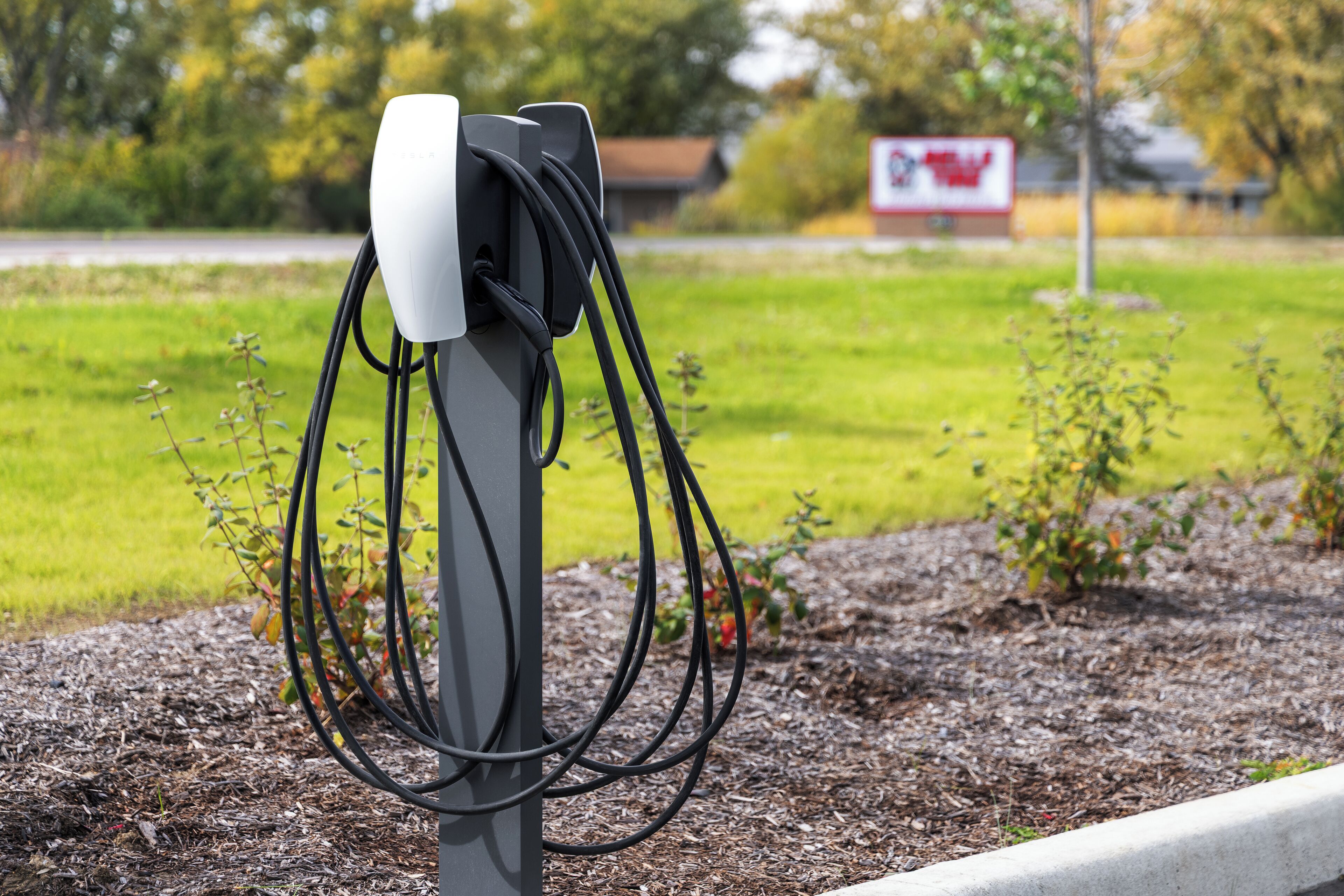 Une borne de recharge pour véhicules électriques, avec des câbles qui y sont raccordés, se dressant sur un fond composé d’une zone herbeuse et d’un bâtiment portant une enseigne.
