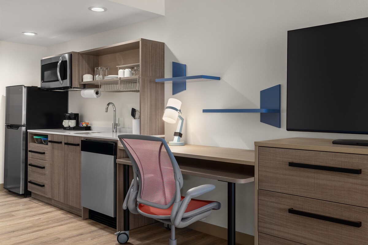 Una cocina moderna con microondas integrado, refrigerador y lavavajillas. Hay un escritorio de madera con una silla, una lámpara de pared y un televisor de pantalla plana.