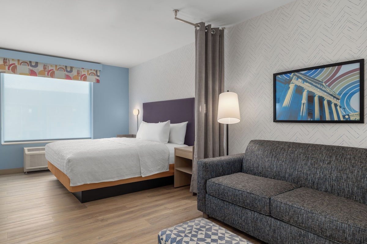 A hotel room with a bed, a sofa, a wall-mounted lamp, and a framed picture.