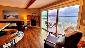 A cozy living room with a fireplace, a dining area, and a balcony with a view of the water. - Lakefront 1 bdrm—Pool & Hot Tub—Lake Michigan Access—2 Kayaks Provided! (Onekama)
