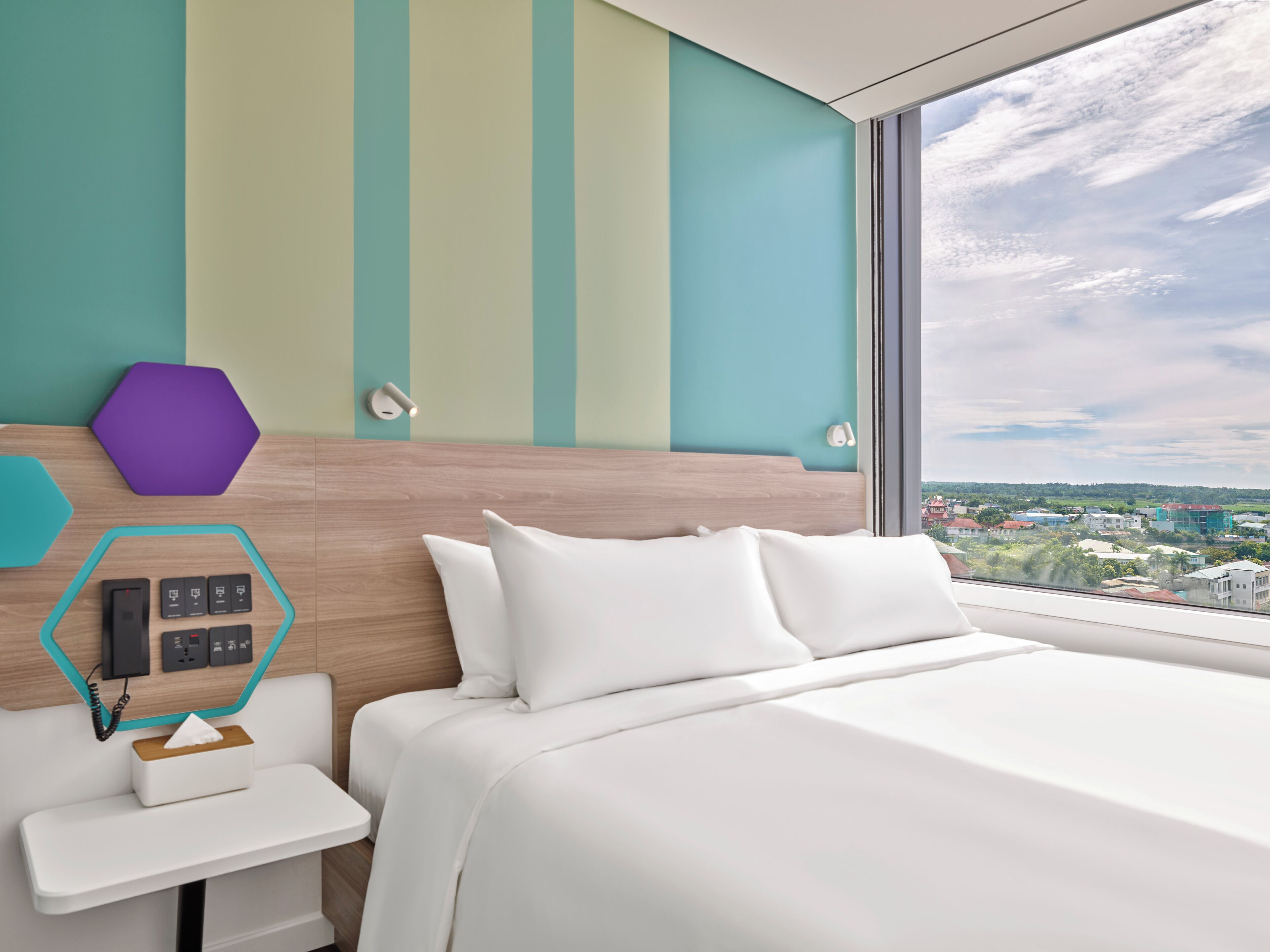 Una habitación de hotel moderna con una cama grande, una mesita de noche pequeña, un panel de control en la pared y un ventanal con vistas a la ciudad.