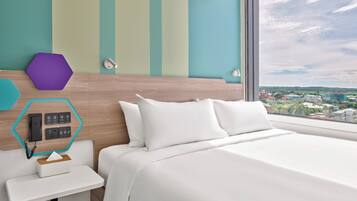 Una habitación de hotel moderna con una cama grande, una mesita de noche pequeña, un panel de control en la pared y un ventanal con vistas a la ciudad.
