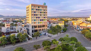 A modern hotel with a prominent logo, surrounded by a cityscape with a clear view of the mountains in the background. - Tru by Hilton Tam Ky City Centre (Da Nang)