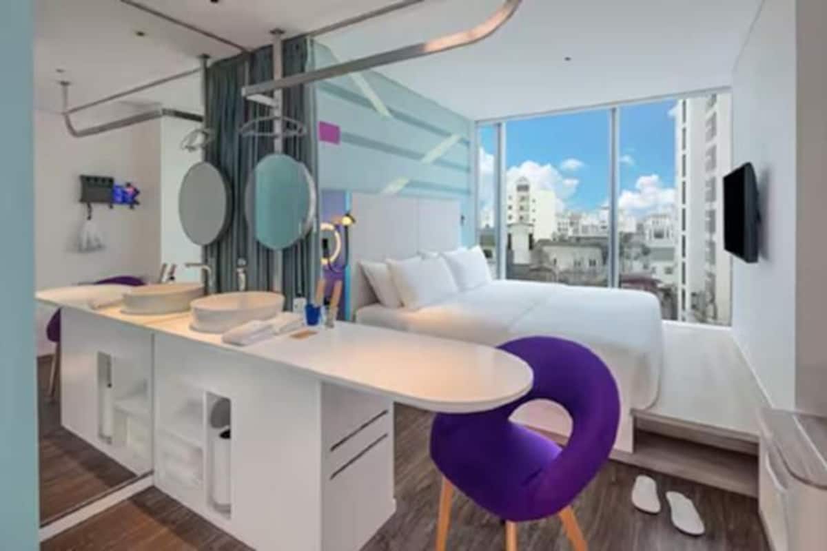 A modern hotel room with a bed, a small sitting area with a table and chairs, a large window with a city view, and a mounted TV.