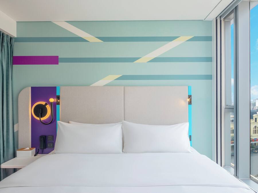 A modern hotel room with a large bed, a headboard with a circular light, and a city view through the window.