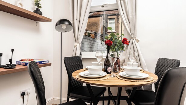 A dining area with a round wooden table set for two, black chairs, a vase with red flowers, and a shelf with books and a plant. - Modern 4bedroom City Home Near LFC (Liverpool)