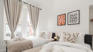 A hotel room with two beds, beige bedding, a nightstand with a lamp, and two framed artworks on the wall. - Modern 4bedroom City Home Near LFC (Liverpool)