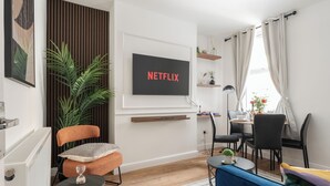A modern living room with a flat-screen TV displaying "NETFLIX", a comfortable orange chair, a blue sofa, and a dining area with a table and chairs. - Modern 4bedroom City Home Near LFC (Liverpool)