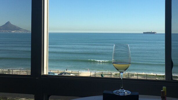 A glass of white wine on a table by a window overlooking a beach with a ship in the distance and a mountain in the background. - Sea View Zeezicht, Bloubergstrand, Blouberg Beach, Cape Town (Bloubergstrand)