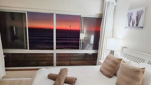 A hotel room with a large window offering a sunset view, a neatly made bed with beige pillows, and a framed picture on the wall. - Sea View Zeezicht, Bloubergstrand, Blouberg Beach, Cape Town (Bloubergstrand)