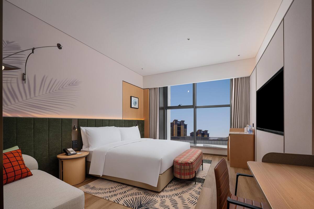 A hotel room with a bed, bedside table, lamp, and a large window with a city view.