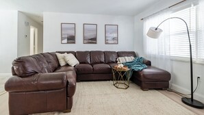 A living room with a brown leather sectional sofa, a small round coffee table, and a floor lamp. There are three framed pictures on the wall and a window with blinds.