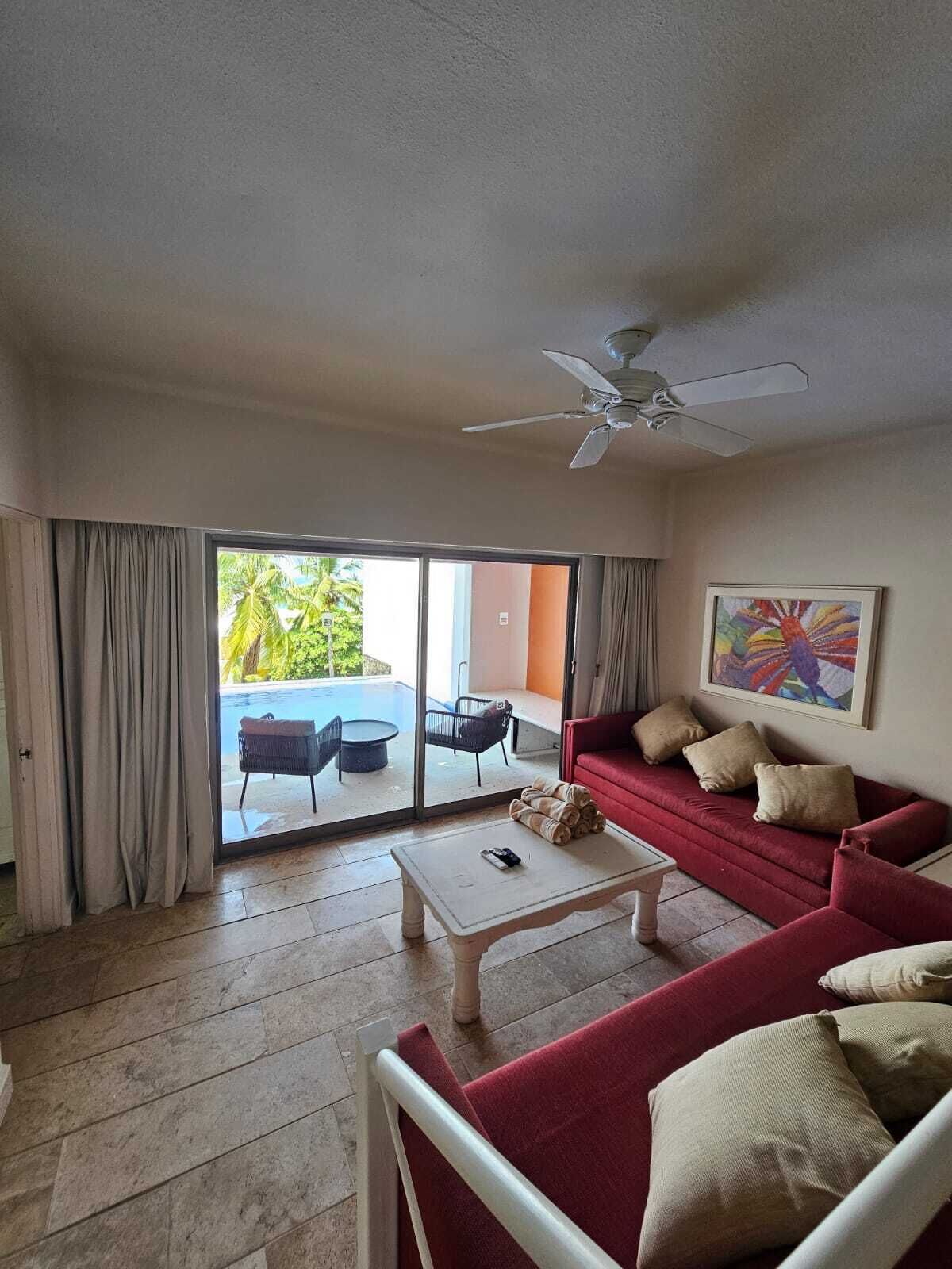 A living room with a red sofa, a white coffee table, and a ceiling fan. There is a sliding glass door leading to an outdoor area with a pool and chairs.