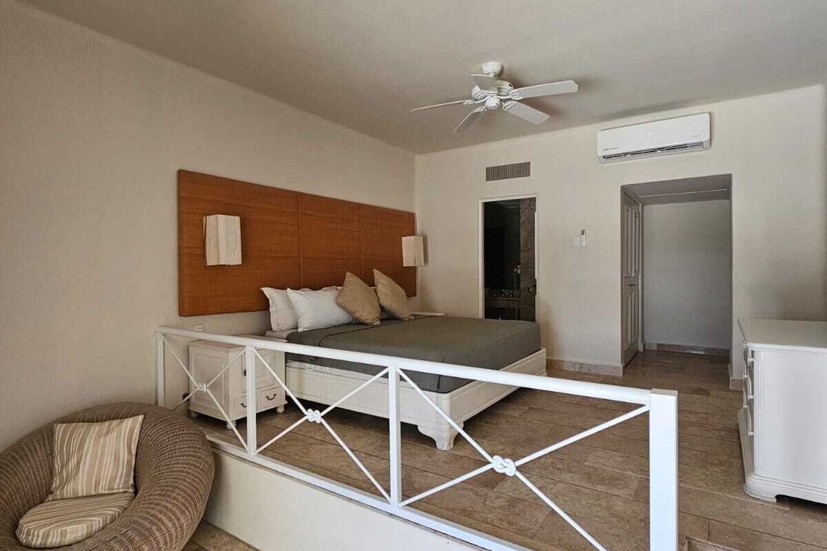Une chambre à coucher comprenant un lit, un ventilateur de plafond, un appareil de climatisation, une armoire blanche et une chaise en osier.