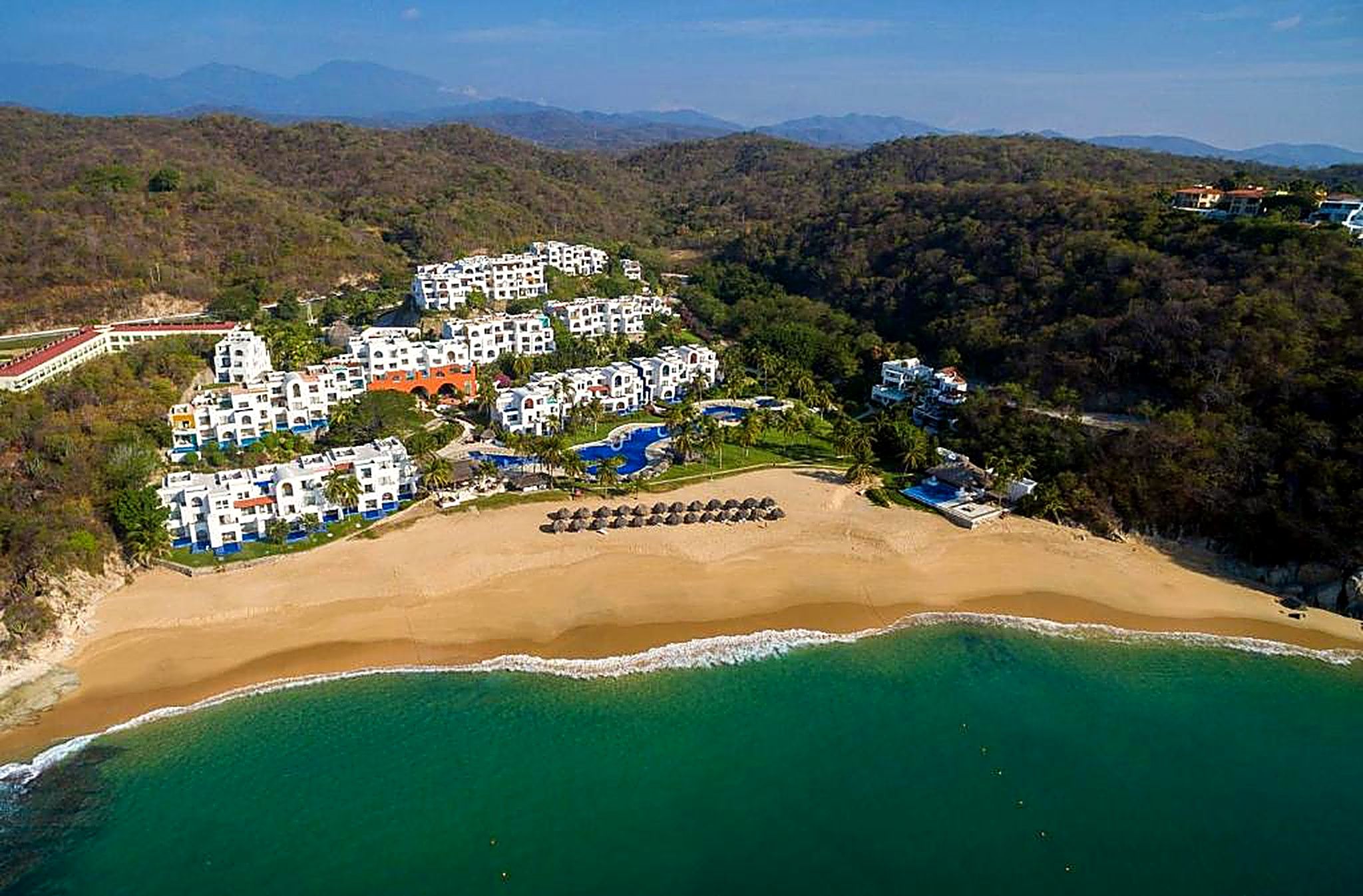 An aerial view of a coastal resort with multiple white buildings, a sandy beach, and a clear blue sea.
