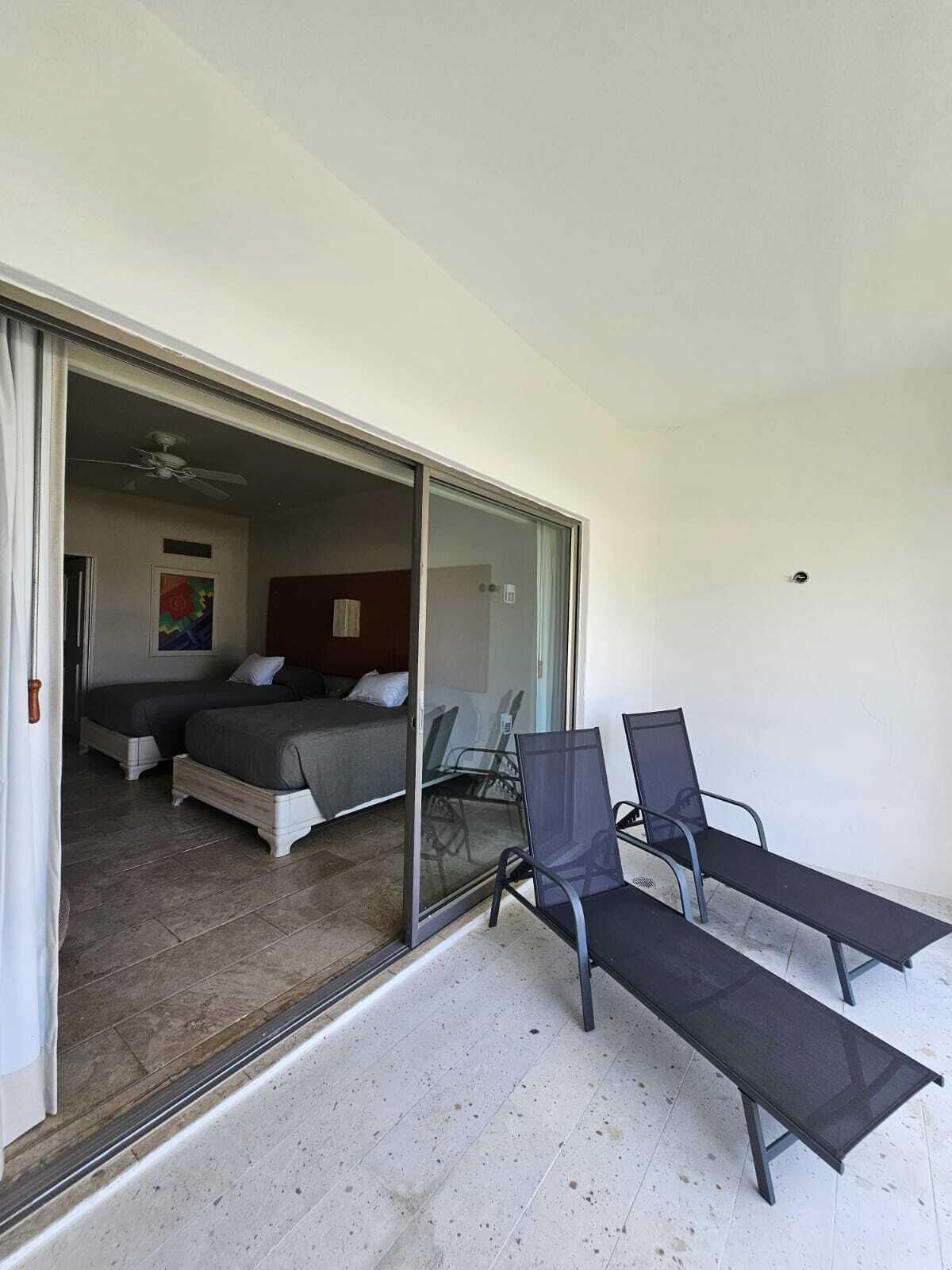 A room with two beds, a sliding glass door leading to an outdoor area with two lounge chairs and a ceiling fan.