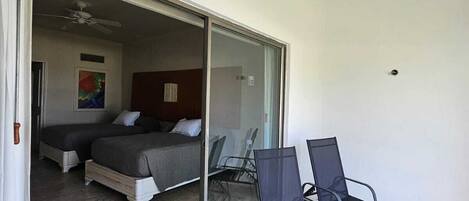 A room with two beds, a sliding glass door leading to an outdoor area with two lounge chairs and a ceiling fan.
