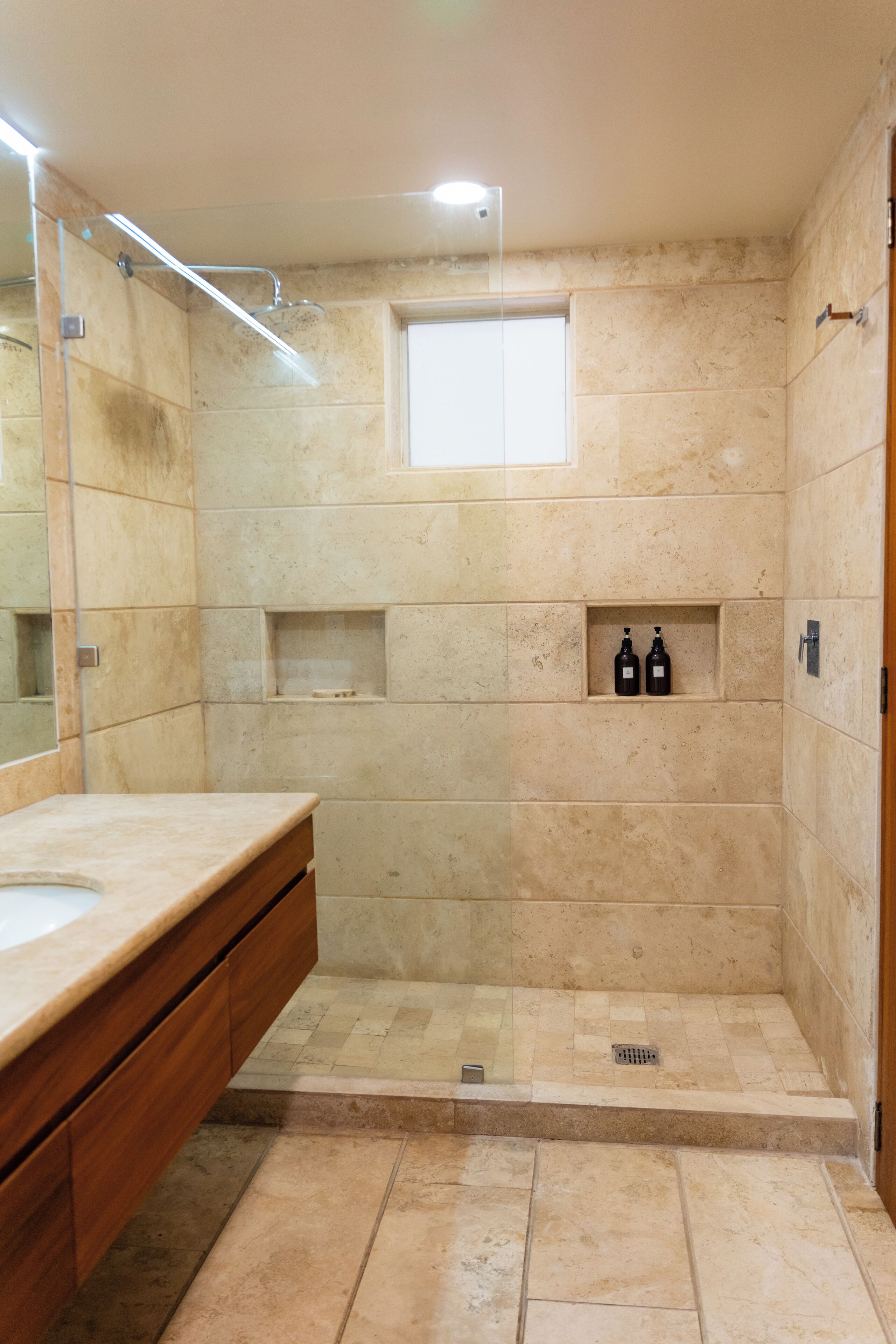 A bathroom with a large mirror, a wooden vanity, a shower area with a glass door, and a small window.