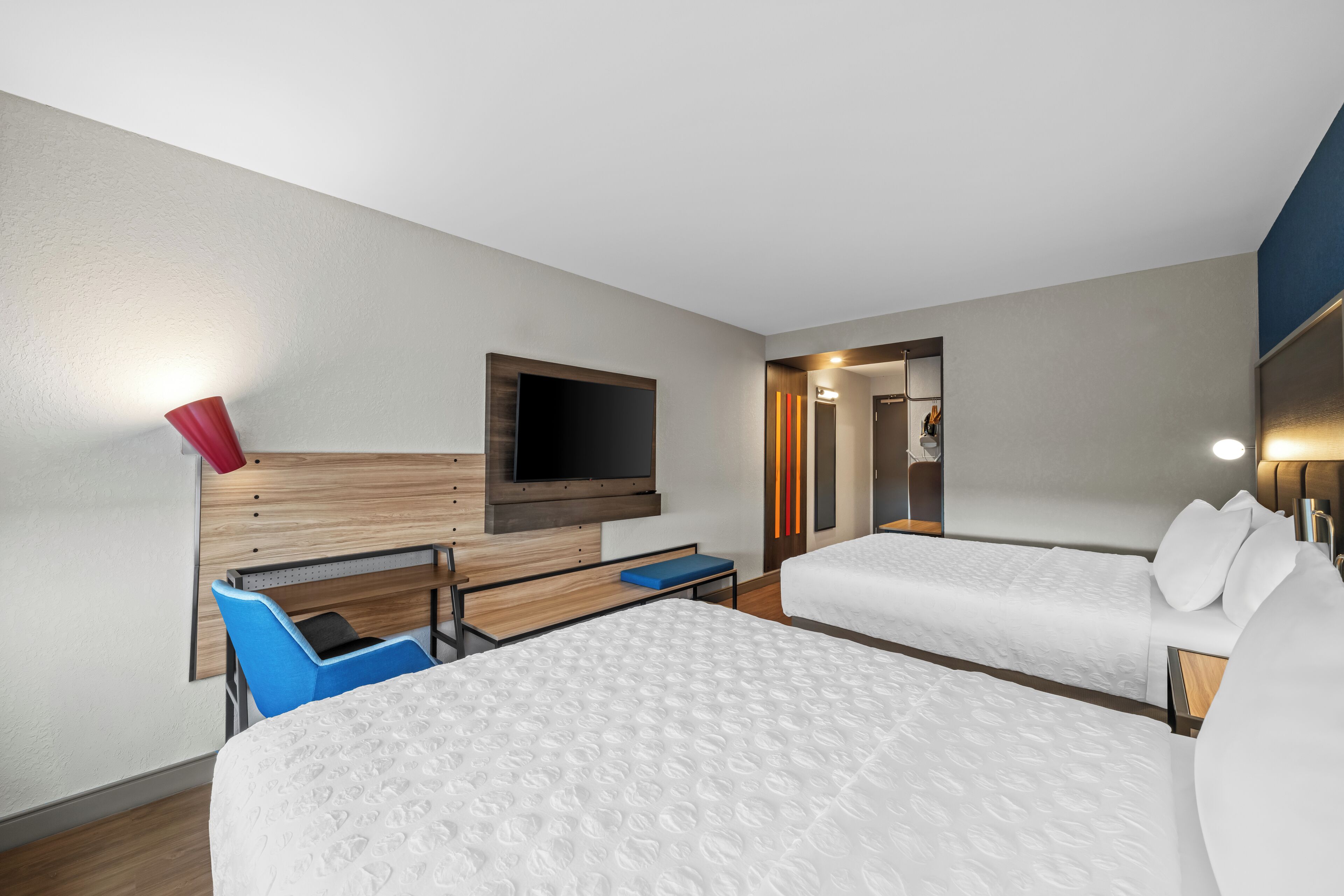 A hotel room with two beds, a TV mounted on the wall, a desk with a chair, and a small table.