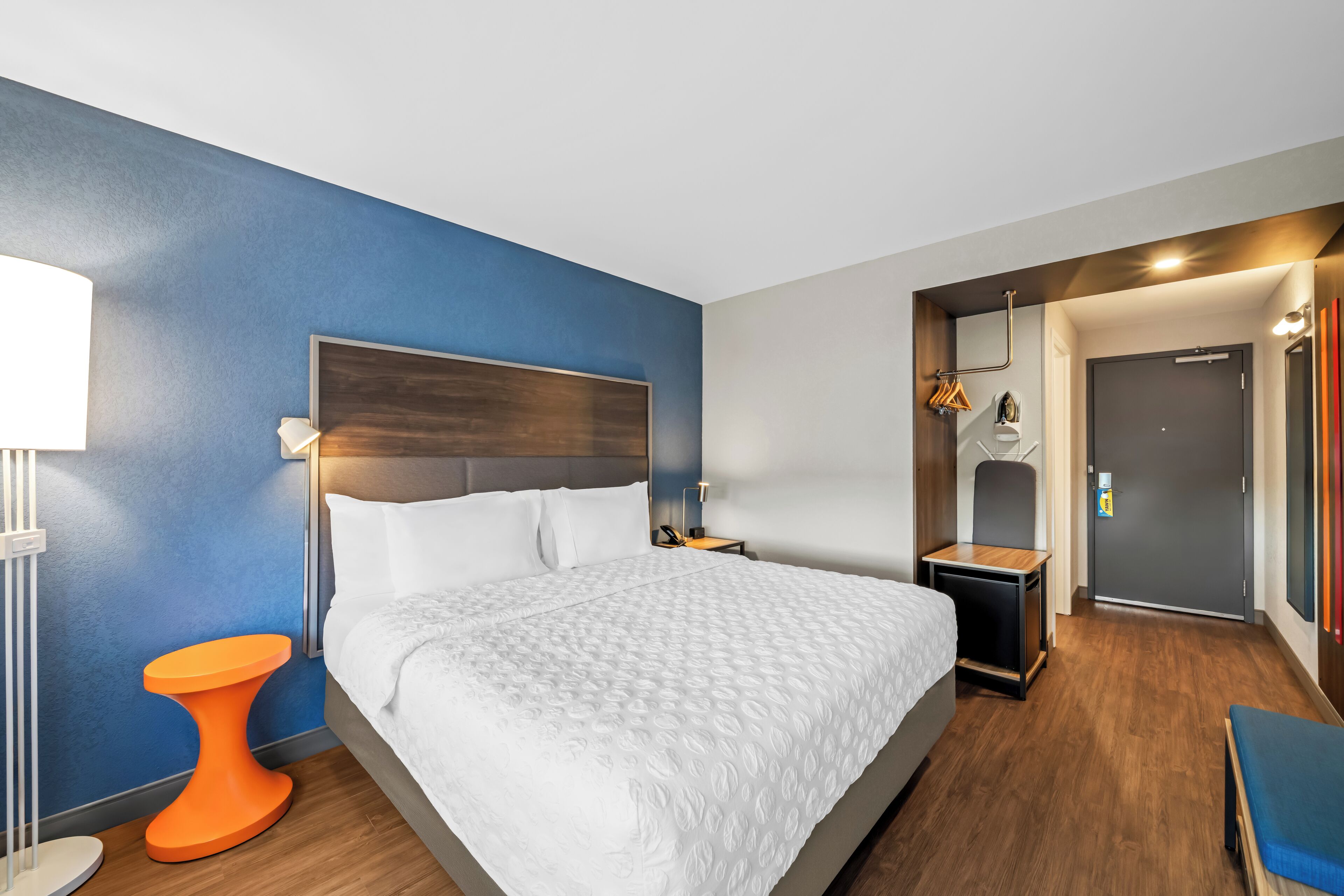 A modern hotel room with a large bed, a small orange side table, a bedside lamp, and a door leading to a bathroom.