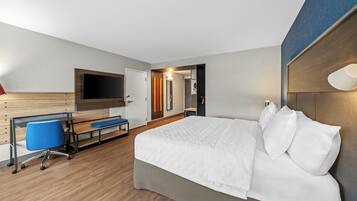 A hotel room with a bed, a desk with a chair, a TV, and a bathroom visible through an open door.