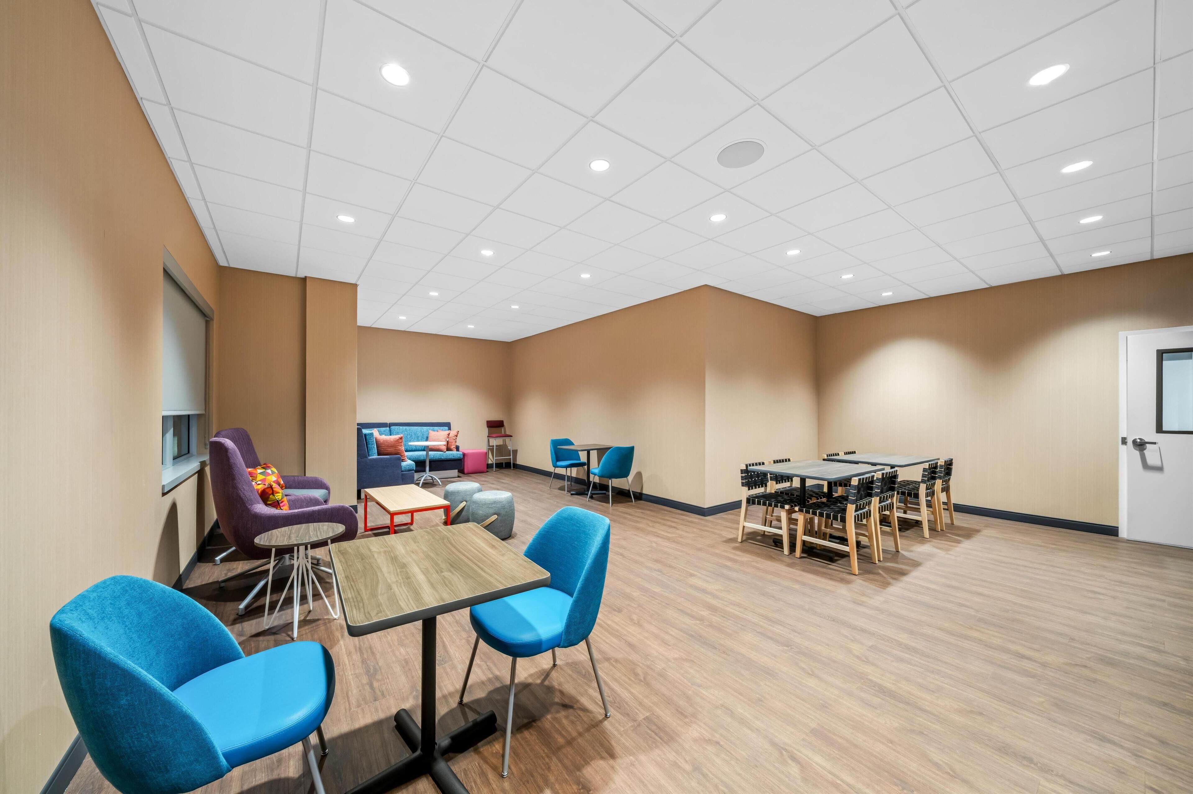 A spacious room with a high ceiling, multiple seating areas including blue chairs and a sofa, a wooden table, and a small kitchenette.