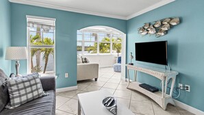 Un salon avec un canapé, un téléviseur et une vue sur les palmiers, visible à travers de grandes fenêtres.