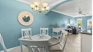 Un coin repas avec une table ronde en verre et des chaises blanches, un lustre, un miroir rond et une vue sur le salon, avec son canapé et sa fenêtre.