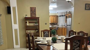 Un comedor con mesa de madera, sillas y una araña. Se ve una cocina al fondo.