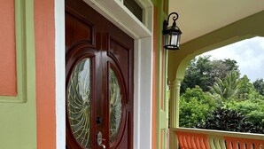 Puertas dobles de madera con un panel de vidrio, un balcón con barandilla decorativa y vistas a la vegetación de fondo.