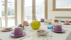 A dining table set with pink and yellow teapots, plates, and cups, with a view of greenery outside through large windows. - The Garden of Aunts - Room of the Heart (Tricase)