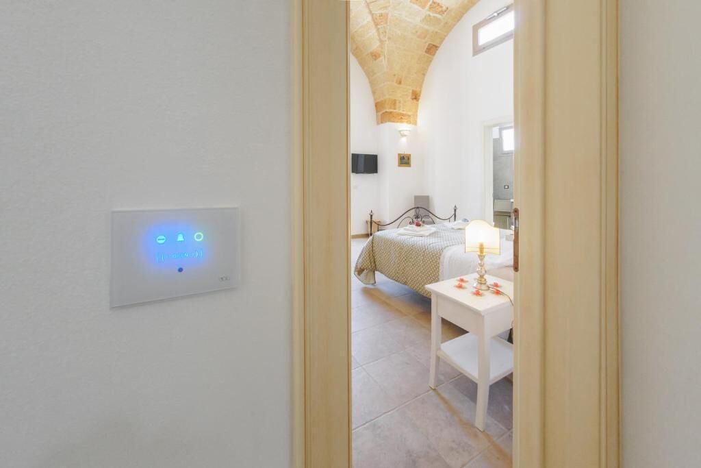 A bedroom with a bed, bedside table, lamp, and a wall-mounted control panel.