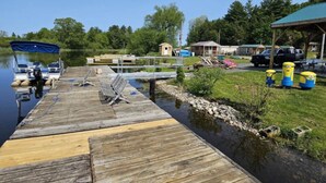 A wooden dock extending into a calm lake, with a small boat docked at the end. There are several buildings and a covered area in the background. - Lovely 1-bedroom cabin on Chippewa Bay, in Hammond, NY (Hammond)