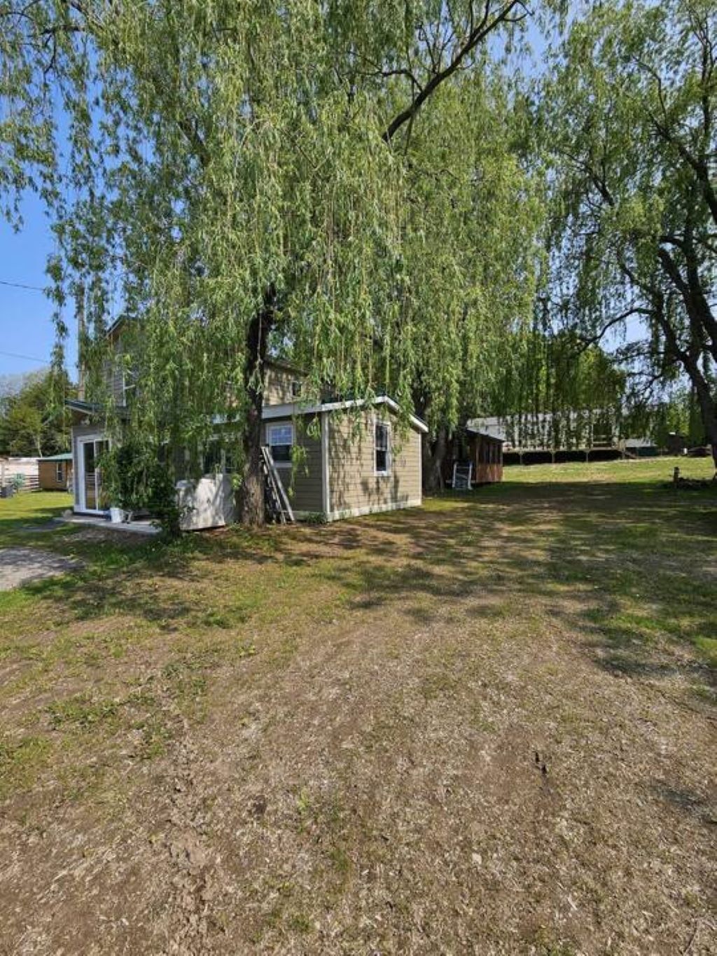 A small, single-story house with a sloped roof, surrounded by a grassy yard and large trees.